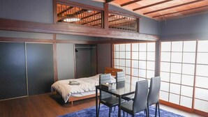 A room with a bed, a dining table with chairs, a large window, and a wooden ceiling.