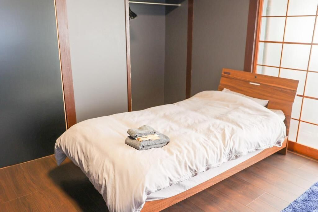 A single bed with a wooden headboard, a grey towel, and a white bedsheet in a room with wooden flooring and a sliding door closet.