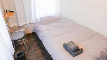 Una habitación pequeña con cama individual, colcha gris, una mesita con silla, un calentador y un cubo de basura.