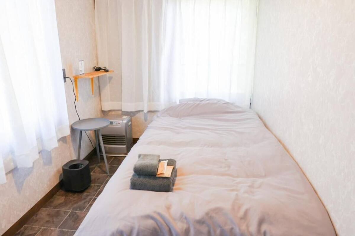 Et lille enkeltværelse med seng, et lille bord, en varmeblæser og en hylde med strømstik.