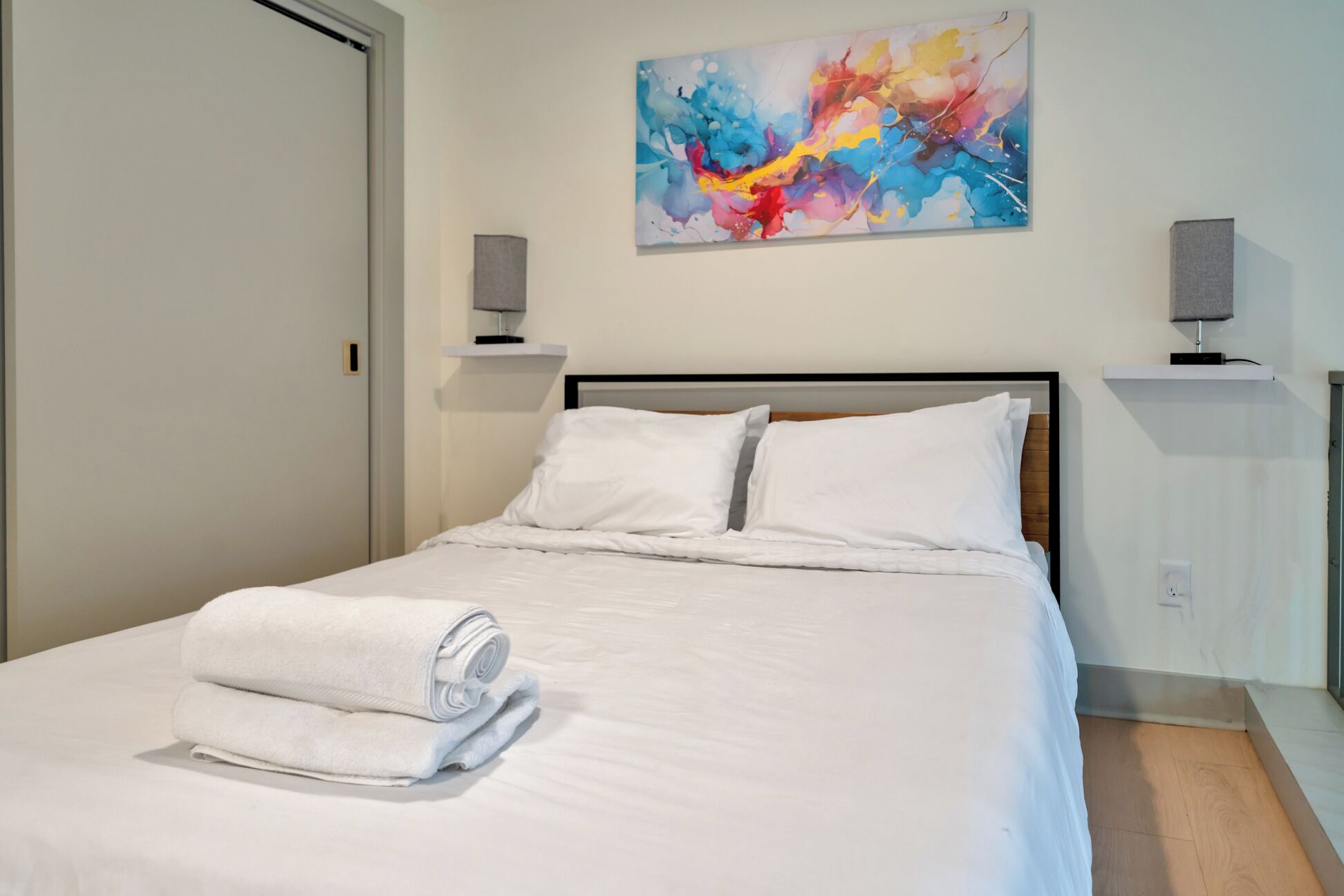 Un lit bien fait, avec du linge de lit blanc et une serviette enroulée, une peinture abstraite aux couleurs vives accrochée au mur, et deux haut-parleurs fixés au mur.