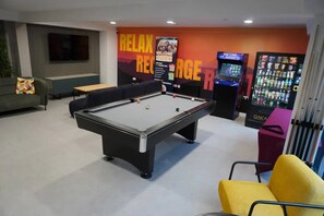 A pool table in a room with a sofa, a vending machine, and a wall with a 'RELAX' sign.