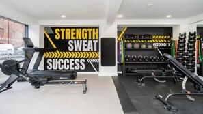 Un gimnasio con equipo de ejercicio, decoración motivacional en la pared y vista a la calle.