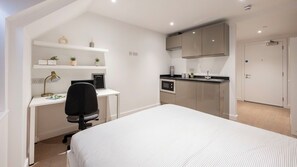Un dormitorio con cama, un escritorio y una silla, una kitchenette y una puerta.
