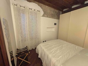 Quarto com uma cama de solteiro, piso de madeira, cortina branca e uma cadeira de madeira pequena.