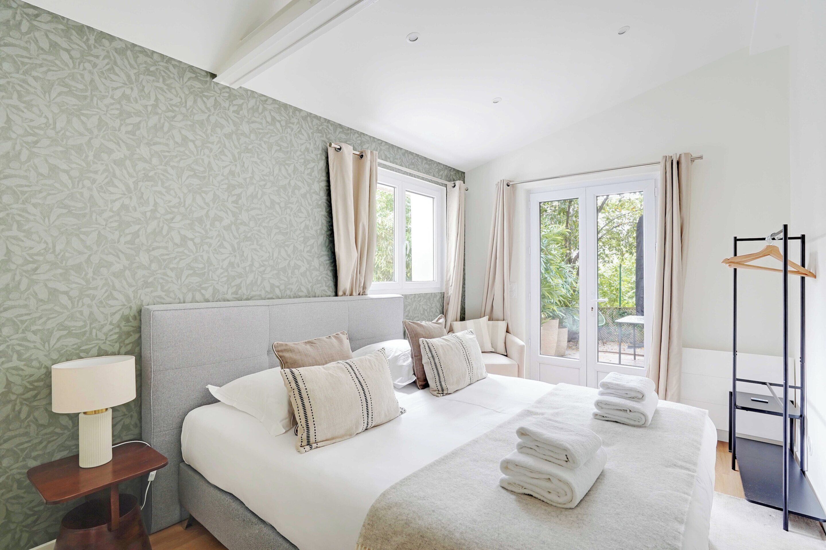 A modern bedroom with a large bed, a bedside table, a lamp, and a view of the outdoors through a window.