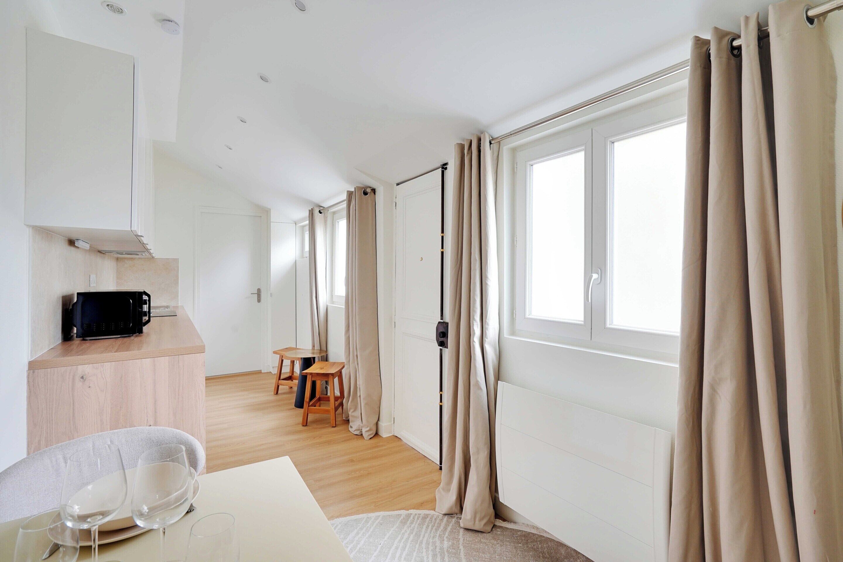 Een moderne, goed verlichte kamer met een keukenhoek, een eettafel met stoelen en grote ramen voorzien van gordijnen.