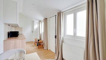 Een moderne, goed verlichte kamer met een keukenhoek, een eettafel met stoelen en grote ramen voorzien van gordijnen.