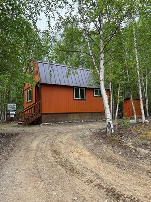 A rustic cabin with a metal roof, wooden deck, and a dirt path leading to it. - Retreat to this quiet 1 bedroom cabin in the woods near a lake. (Wasilla)