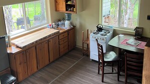 A kitchen with wooden cabinets, a stove, and a dining area with chairs. - Retreat to this quiet 1 bedroom cabin in the woods near a lake. (Wasilla)