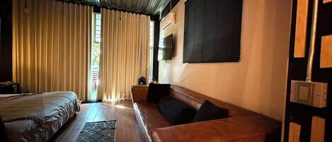 A room with a bed, a sofa, a television mounted on the wall, and a window with curtains.