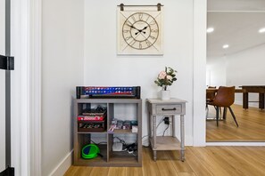 Ein Zimmer mit Holzboden, einer Wanduhr, einem Regal mit Gesellschaftsspielen, einem kleinen Tisch mit einer Vase und einem Essbereich im Hintergrund.