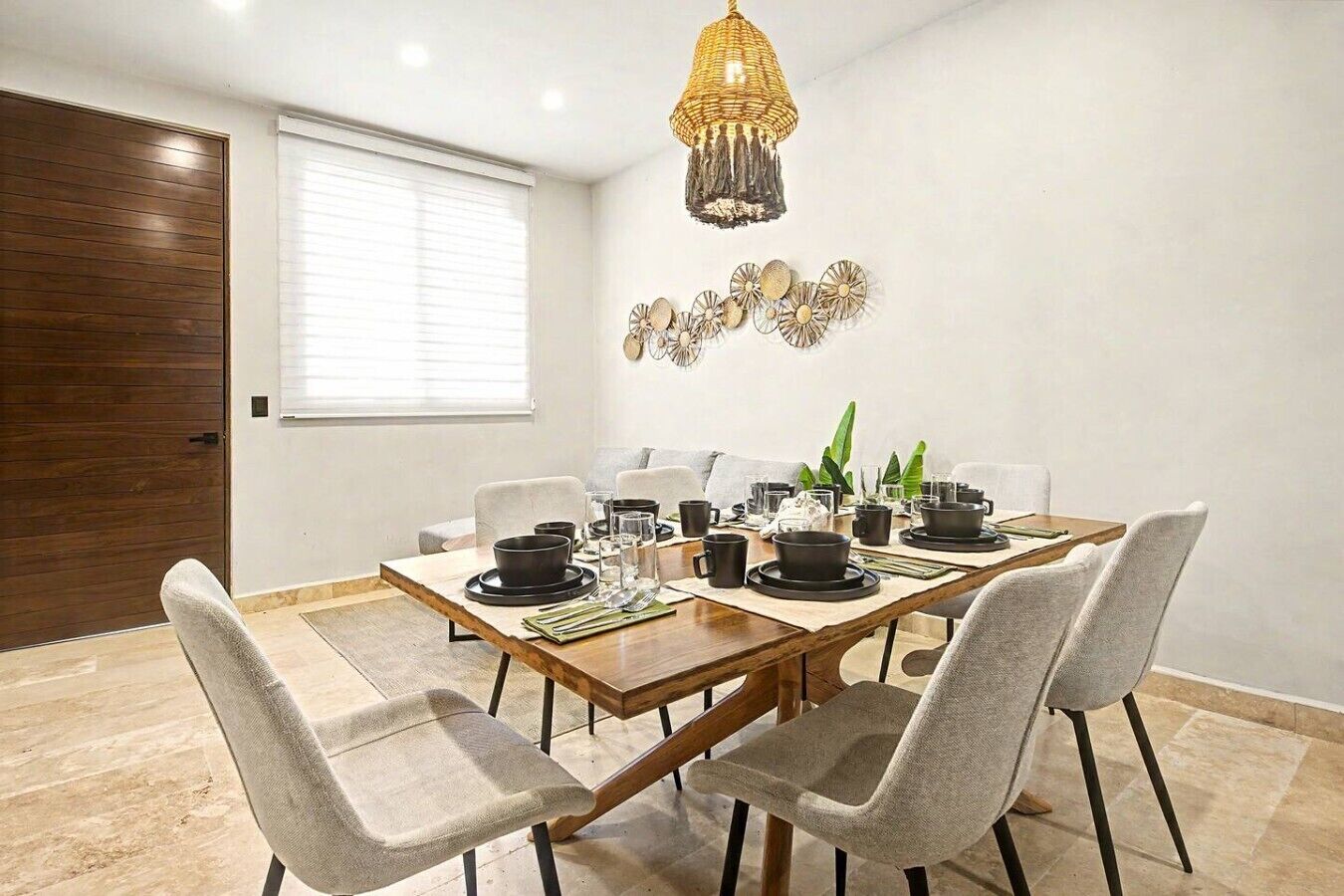 A dining area with a wooden table set for four, surrounded by grey chairs, a hanging light fixture, and a wall-mounted decorative piece.