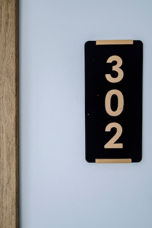 Letrero de número de habitación en color negro y dorado con los números '302' sobre una pared blanca.