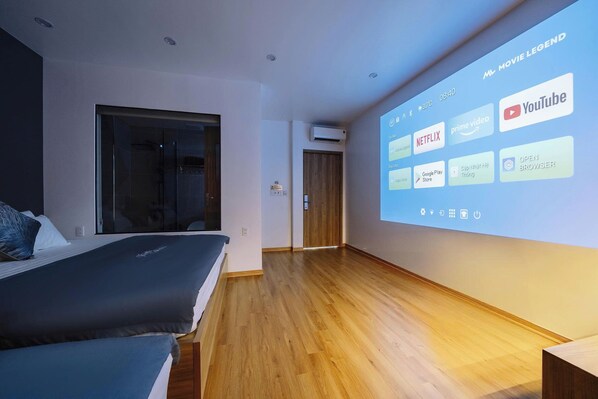A modern hotel room with a large bed, a small desk, and a wall-mounted projector displaying various streaming services.