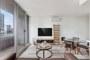 A modern living room with a large TV, a dining area with a glass table, and a view of the city through large windows. - La Trobe Lookout Poolside Retreat by State Library (Melbourne)