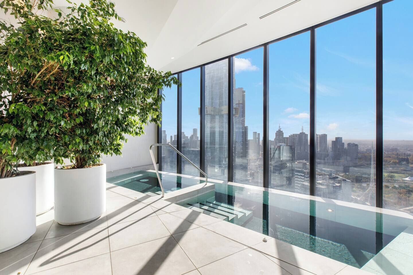 A modern indoor pool area with a city view, large potted plants, and a glass barrier.