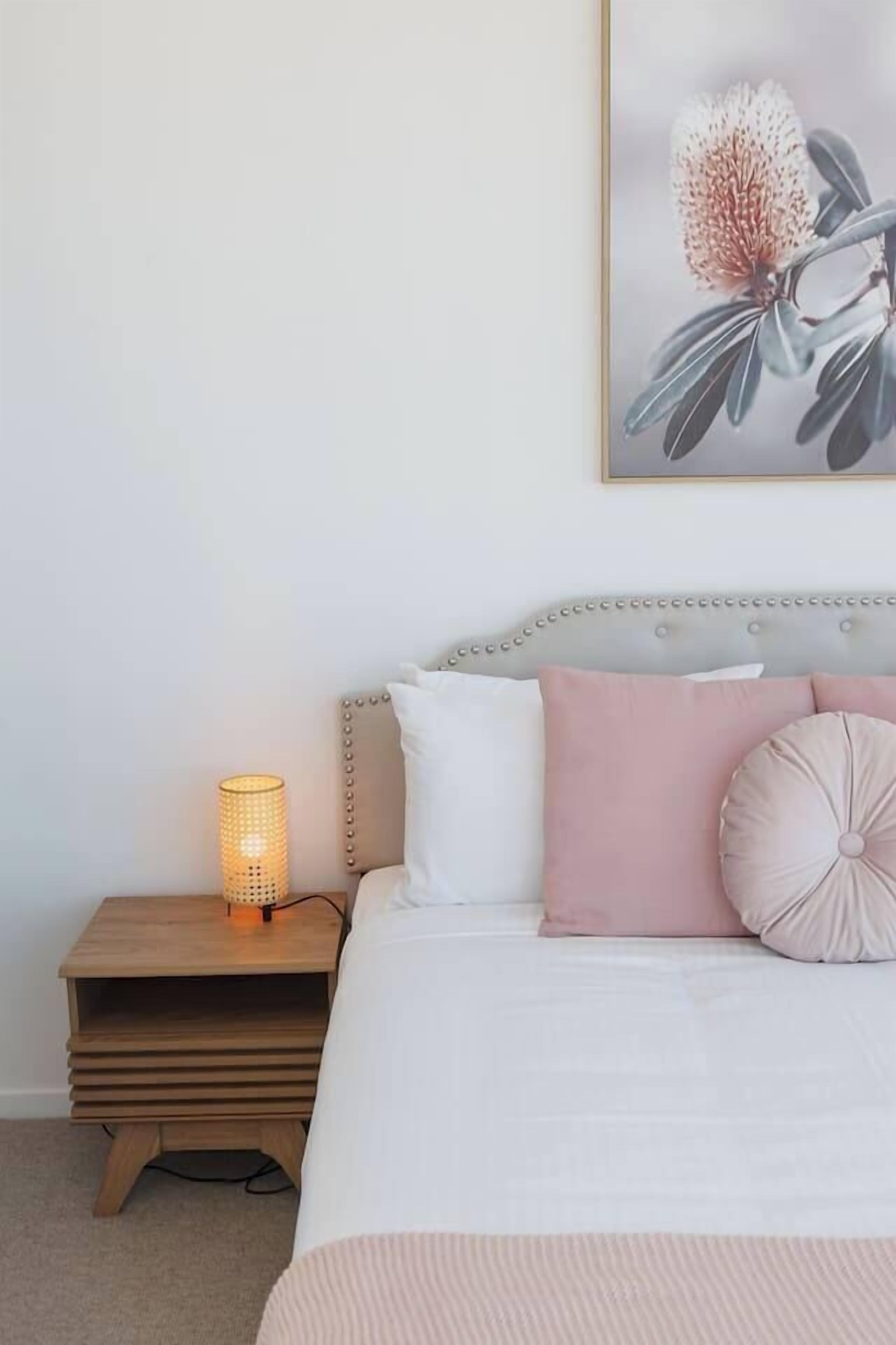 Une chambre à coucher avec un lit, des oreillers, une lampe de chevet et une œuvre d’art florale encadrée accrochée au mur.