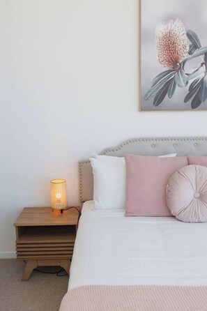 Ein Schlafzimmer mit einem Bett, Kissen, einer Nachttischlampe und einem Blumenbild in einem Rahmen an der Wand.