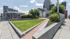 Jardim no terraço com um caminho pavimentado, cercado por edifícios modernos e um céu claro.