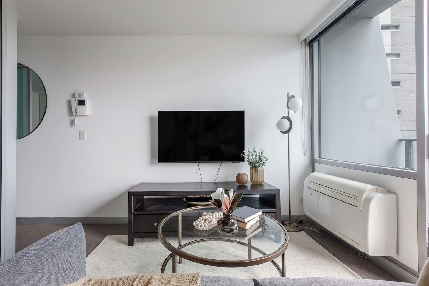 Un salon moderne avec un téléviseur à écran plat fixé au mur, un meuble de rangement en bois foncé, une table basse en verre agrémentée d’objets décoratifs et une grande fenêtre offrant une vue sur l’extérieur.