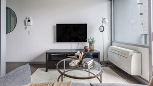 Uma sala de estar moderna com televisão de tela plana na parede, um móvel de entretenimento em madeira escura, uma mesa de centro de vidro com objetos decorativos e uma grande janela com vista para o exterior.