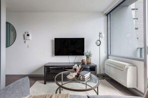 A modern living room with a flat-screen TV mounted on the wall, a dark wooden entertainment unit, a glass coffee table with decorative items, and a large window providing a view of the outside. - Batman Hideout Modern Parkside Escape with Gym (West Melbourne)