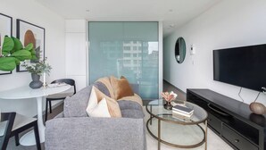 Uma sala de estar moderna com um sofá cinza, uma mesa de centro redonda, uma televisão de tela plana e uma parede divisória de vidro.