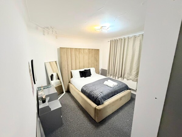 A hotel room with a bed, a bedside table, a mirror, a window with curtains, and a light fixture.