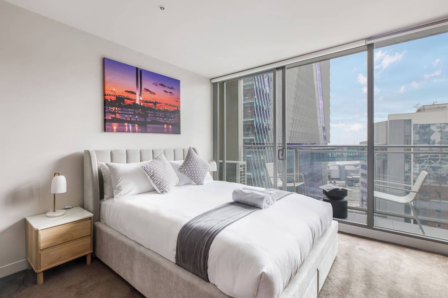 Une chambre d’hôtel moderne avec un grand lit, une table de chevet, une lampe et une vue sur la ville par la fenêtre.