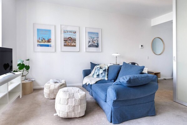 A modern living room with a blue sofa, a white ottoman, and a television on a white stand. There are framed pictures on the wall and a round mirror on the right. - Urban Cocoon Cosy Riverside Pad with Pool and Sauna (Southbank)