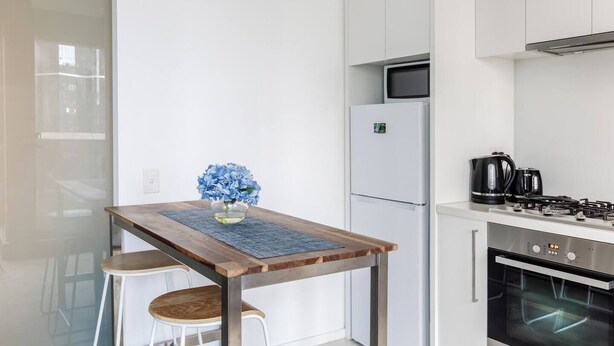 A modern kitchen with a white refrigerator, stove, and oven. There is a wooden table with a vase of flowers and two stools. - Urban Cocoon Cosy Riverside Pad with Pool and Sauna (Southbank)