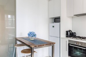 A modern kitchen with a white refrigerator, stove, and oven. There is a wooden table with a vase of flowers and two stools. - Urban Cocoon Cosy Riverside Pad with Pool and Sauna (Southbank)