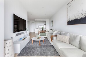 Ein modernes Wohnzimmer mit Flachbildfernseher, einem weißen Sofa, einem Couchtisch und einer Küche im Hintergrund.