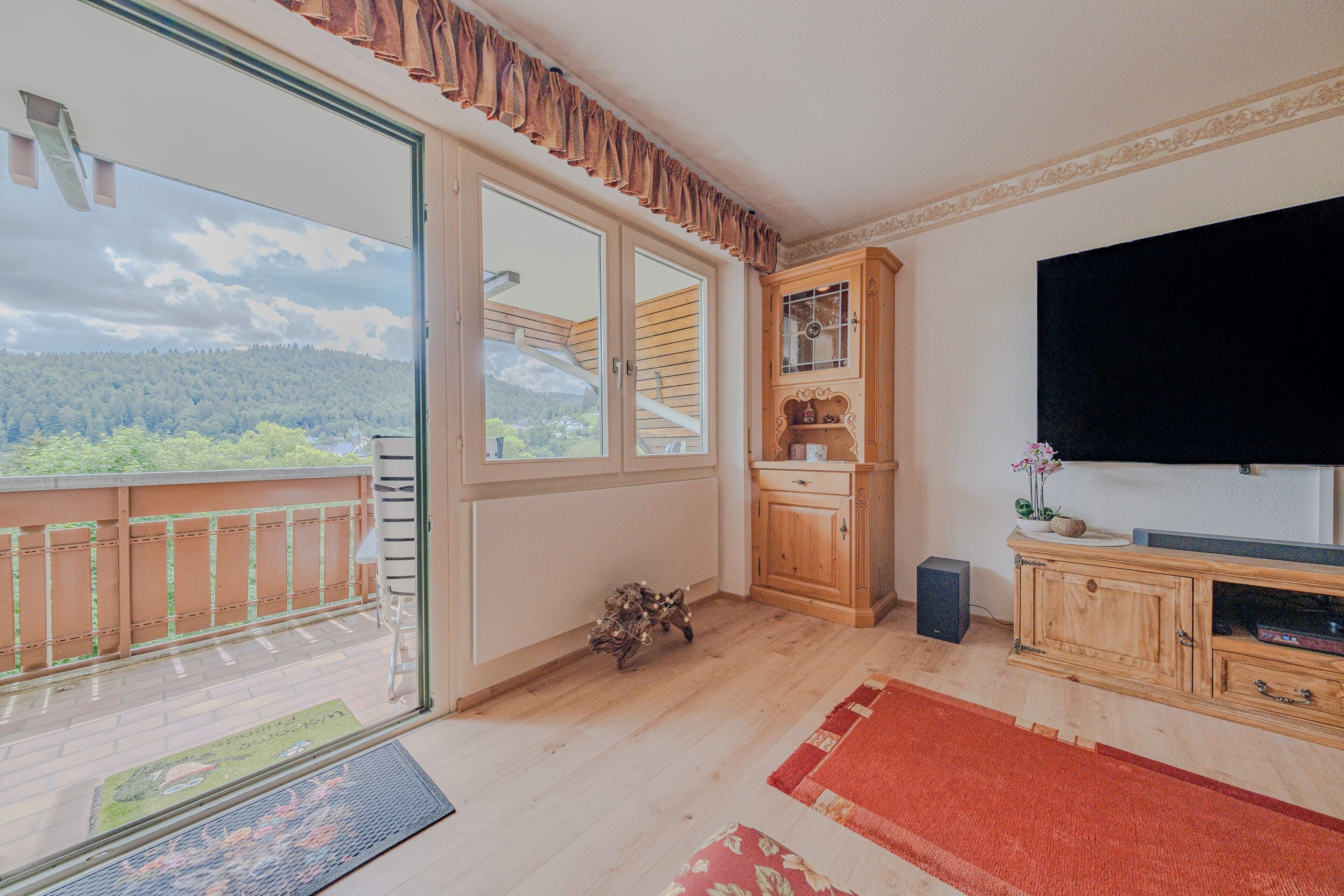 Ein Zimmer mit Balkon, einem Holzschrank, einem Fernseher und Blick auf ein bewaldetes Gebiet.