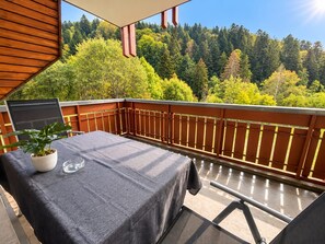 A balcony with a table and chairs, a potted plant, and a view of a forested hillside. - Pine Forest Bliss – A Wonderful Vacation with a Stunning Balcony View – My Little Black Forest Cottage (Todtmoos)