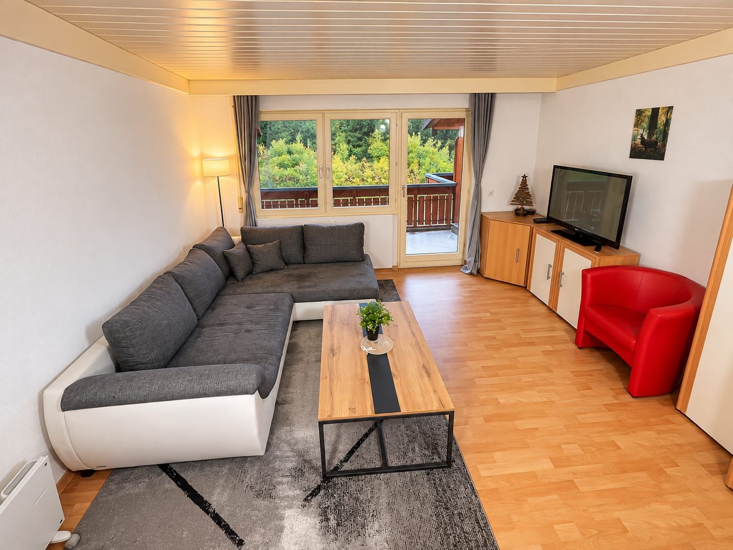 A modern living room with a sectional sofa, a wooden coffee table, a red armchair, a television, and a sliding glass door leading to a balcony.