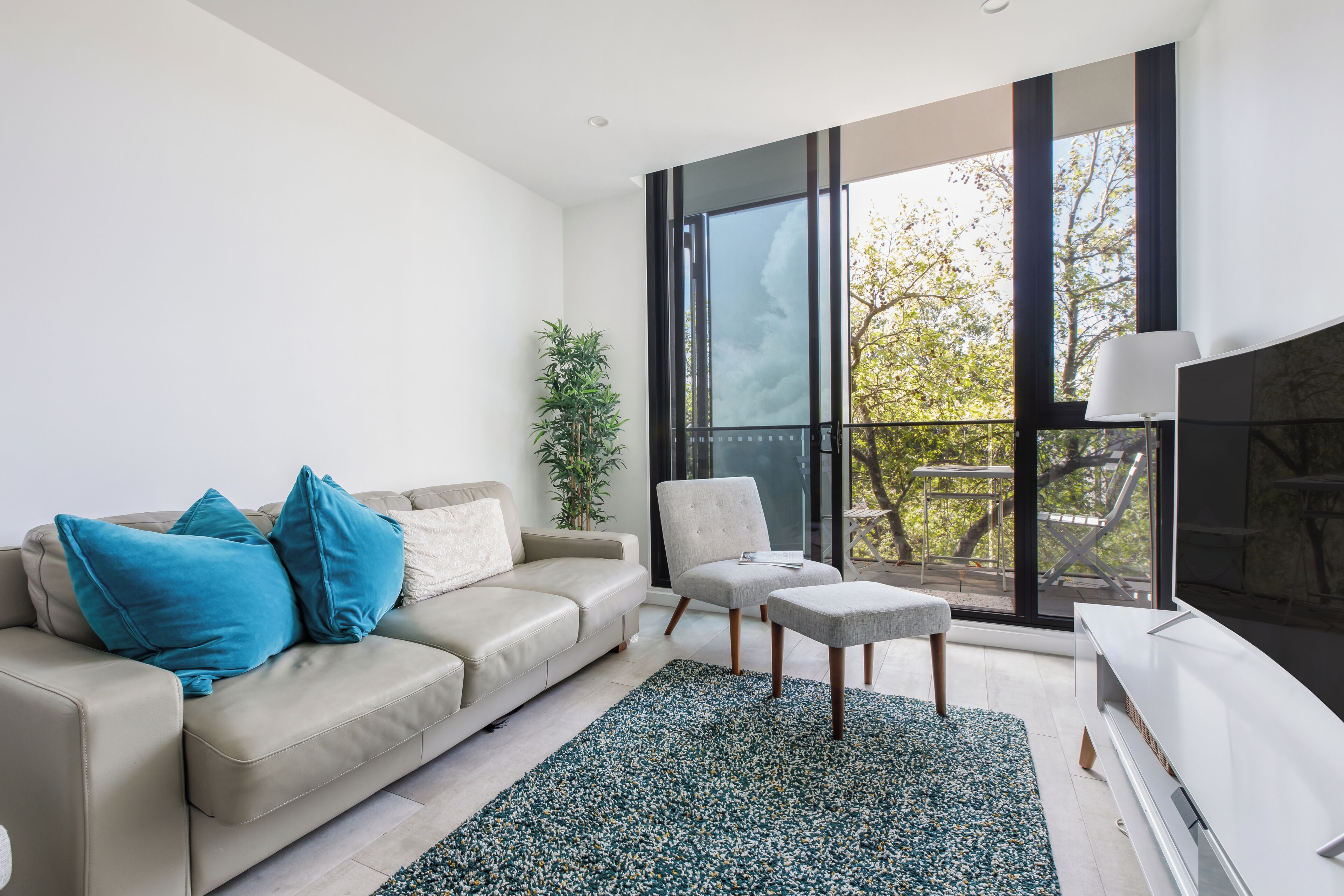 Een moderne woonkamer met een beige bank, blauwe kussens, een grijze fauteuil, een klein poufje en een groot raam met uitzicht op bomen.