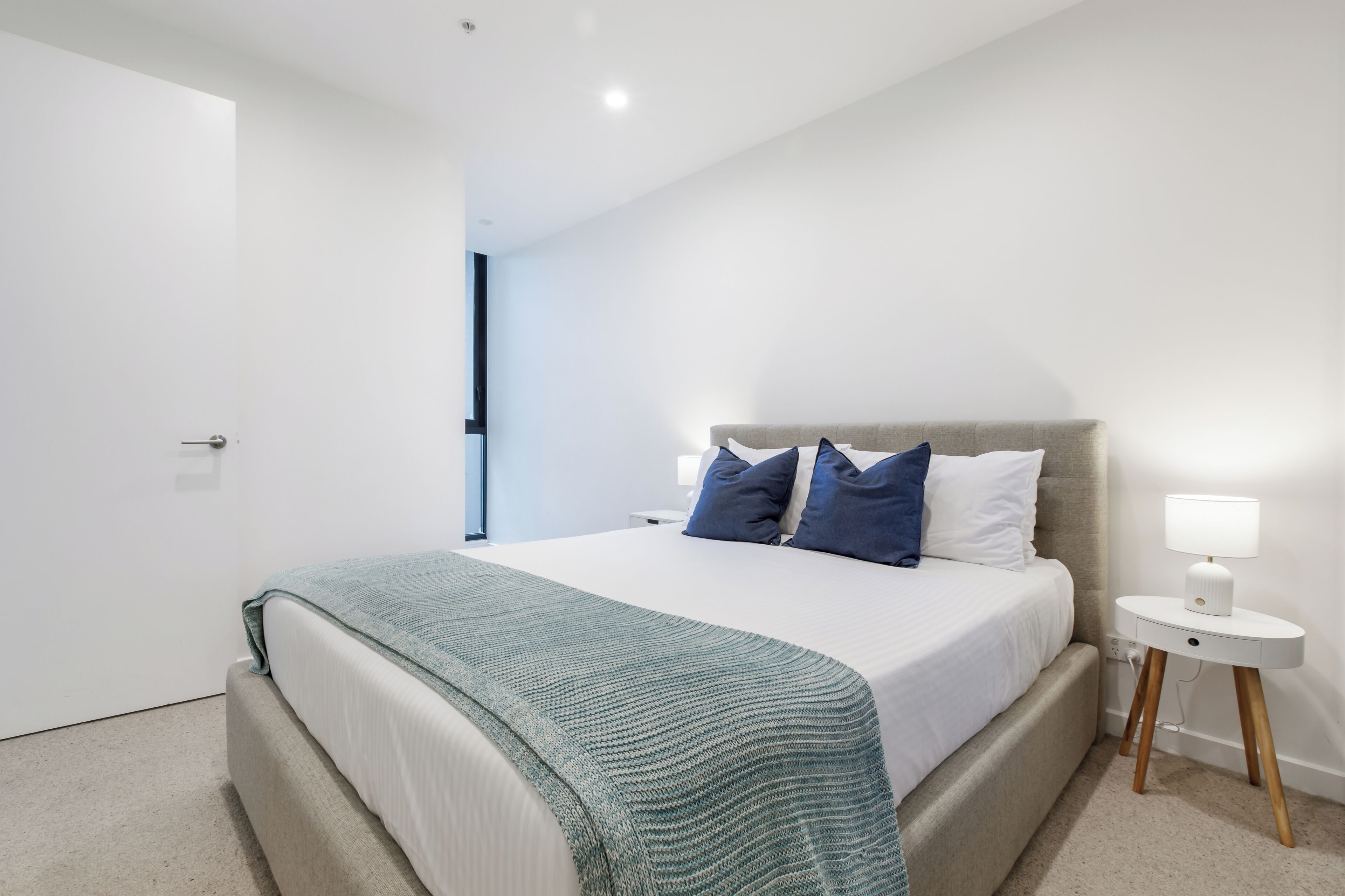 A modern bedroom with a large bed, a bedside table with a lamp, and a view of the hallway.