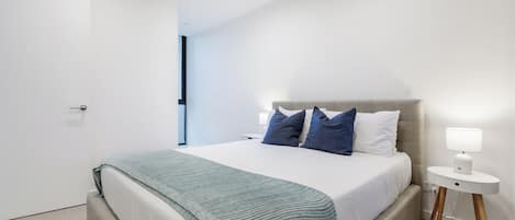 A modern bedroom with a large bed, a bedside table with a lamp, and a view of the hallway.