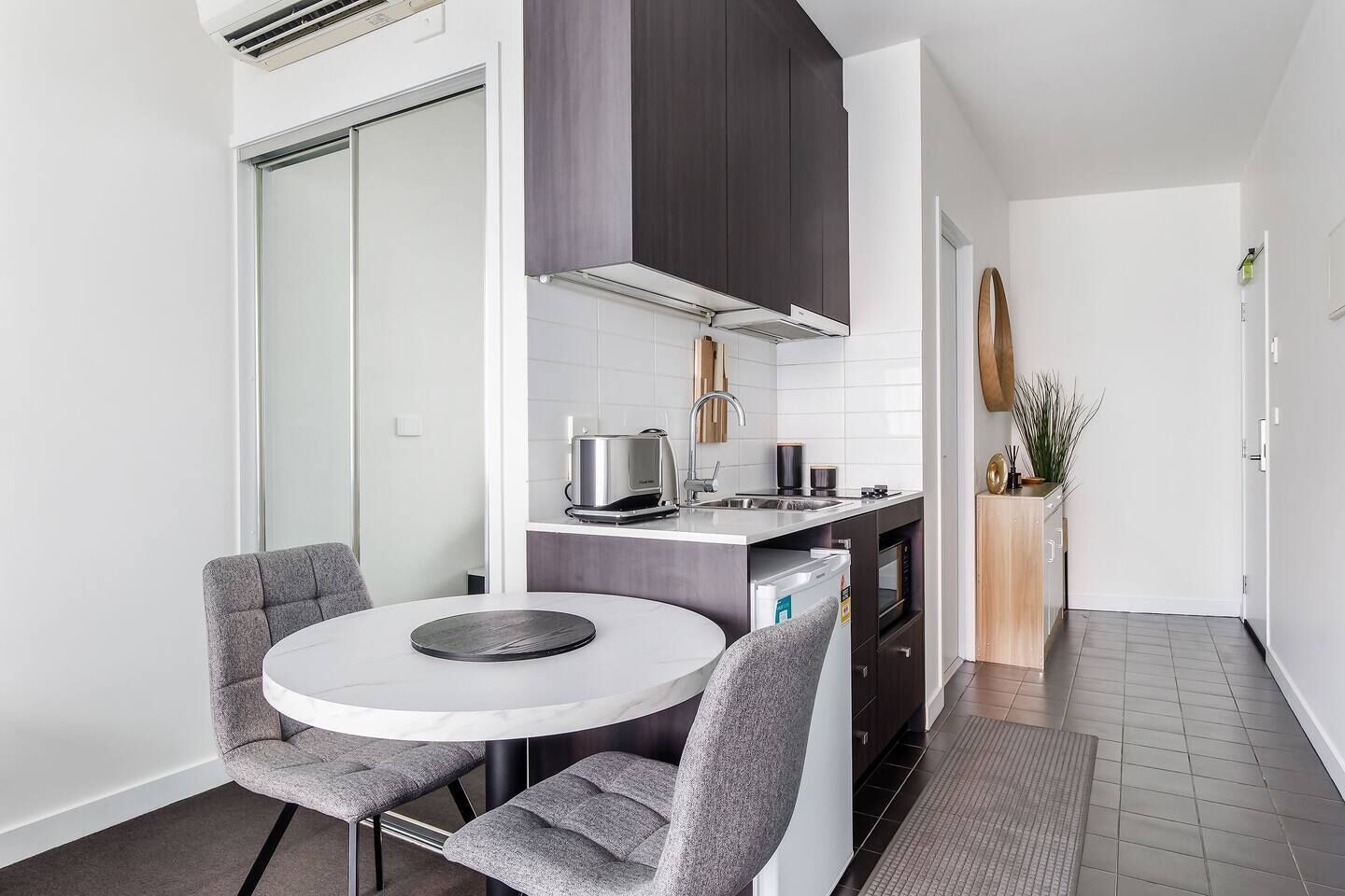 Een compacte keuken met een ronde eettafel, grijze gestoffeerde stoelen, een inbouwoven en een magnetron.