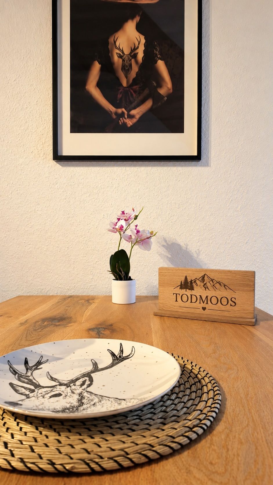 A framed picture on a wall, a small potted plant, a wooden sign, and a plate with a deer illustration on a table.