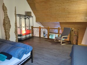 Een kamer met een schuine balkenlaag, een bed met een blauwe deken, een stoel en een klein tafeltje met een lamp.