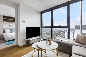 Una sala de estar moderna con un televisor de pantalla plana, un sofá, una mesa de centro y vistas a la ciudad a través de amplios ventanales.