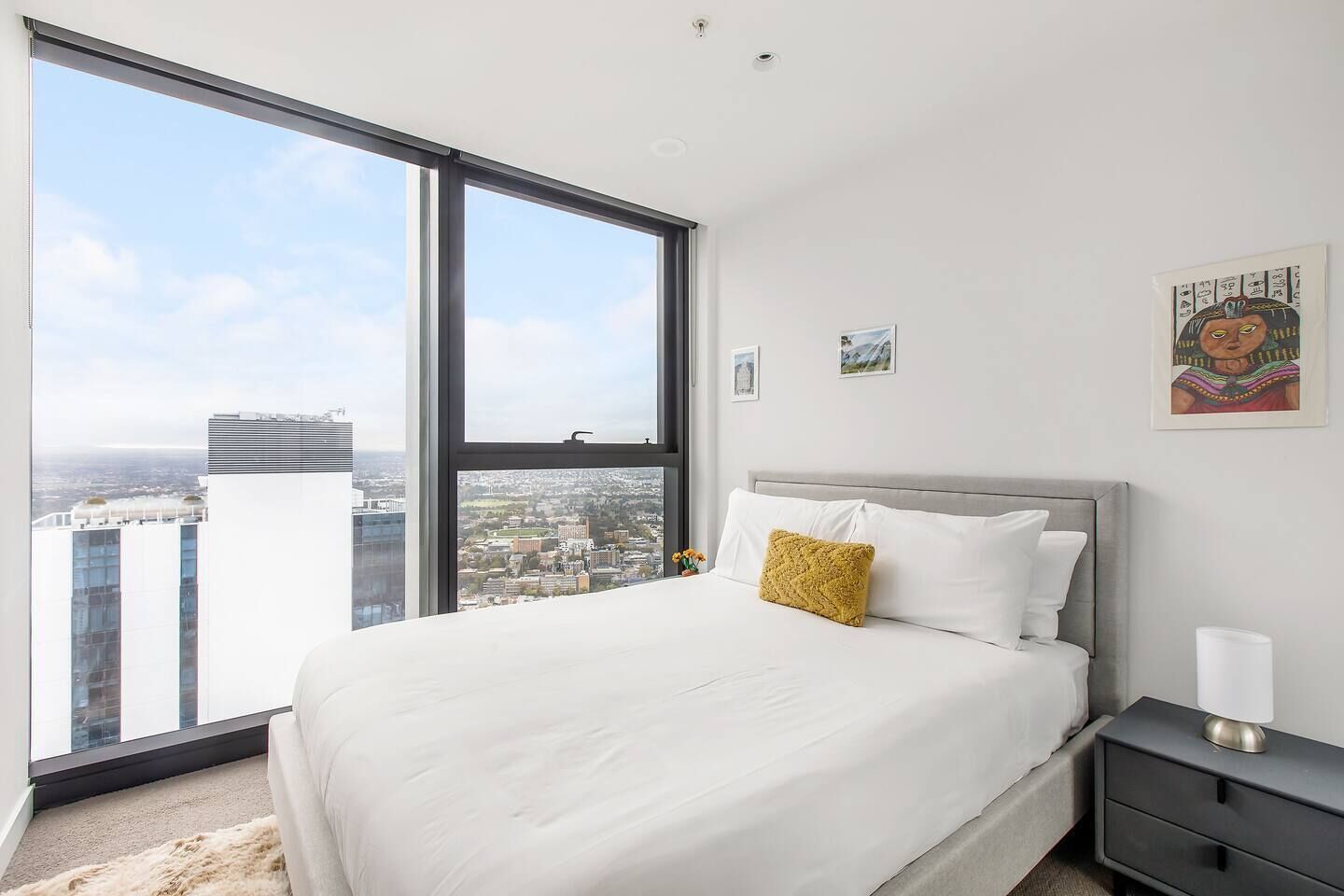 Une chambre avec une grande fenêtre, un lit avec une literie blanche et un oreiller jaune, une table de chevet avec une lampe, et une vue sur le paysage urbain.