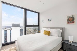 Un dormitorio con un ventanal grande, una cama con ropa de cama blanca y almohada amarilla, una mesita de noche con lámpara, y vista a la ciudad.