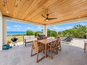 An outdoor patio with a wooden table and chairs, a grill, and a view of the ocean.
