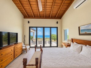 A bedroom with a large bed, a television, a wooden dresser, and a view of the ocean through large sliding doors.