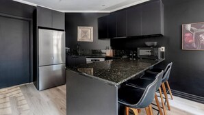 A modern kitchen with dark cabinetry, a stainless steel refrigerator, a granite countertop, and bar stools. - Noir Haven Stylish CBD Escape with Private Rooftop (Melbourne)
