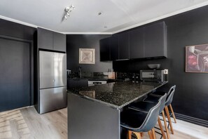 A modern kitchen with dark cabinetry, a stainless steel refrigerator, a granite countertop, and bar stools. - Noir Haven Stylish CBD Escape with Private Rooftop (Melbourne)